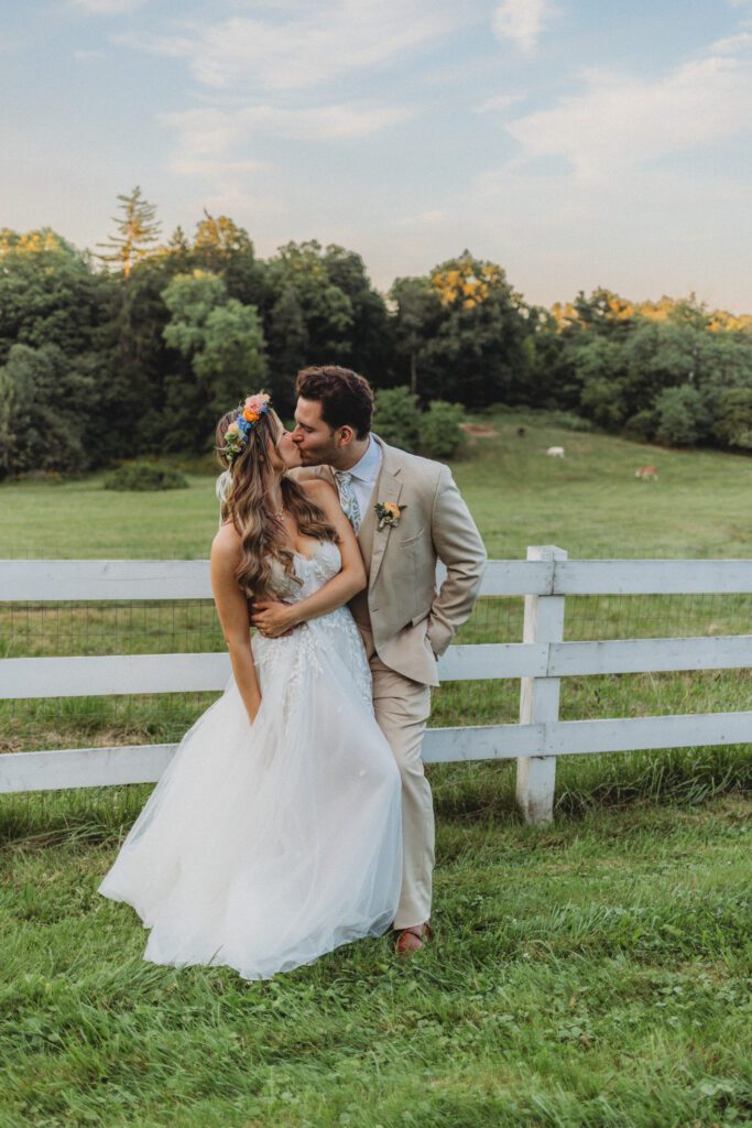 Bride and groom standing front of a white fence kissing at whimsical wedding at Peirce Farm Estate in Topsfield, Massachusetts.