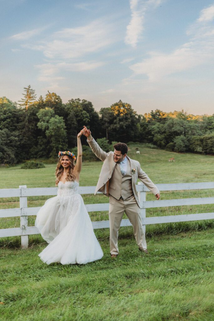Bride and groom laughing and spinning in front of white fence at whimsical wedding Groom romantically spinning bride in front of white fence during sunset at Peirce Farm Estate in Topsfield, Massachusetts.
