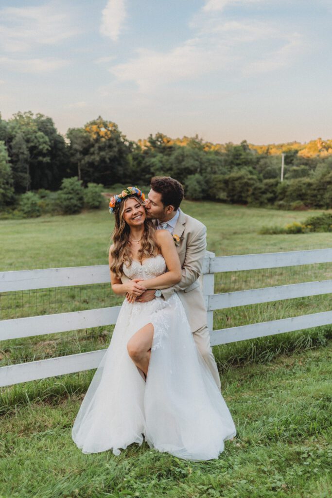 Groom kissing bride on the cheek as they stand in front of a white fence smiling at Peirce Farm Estate in Topsfield, Massachusetts.