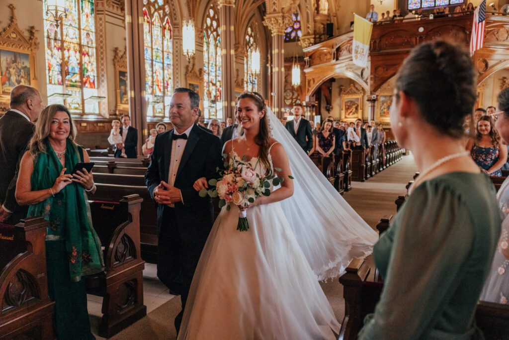 Bride and her father walking down the aisle with the bride veil dragging behind her in church ceremony at Quinet Saint John Garden of Our Lady in Massachusetts.