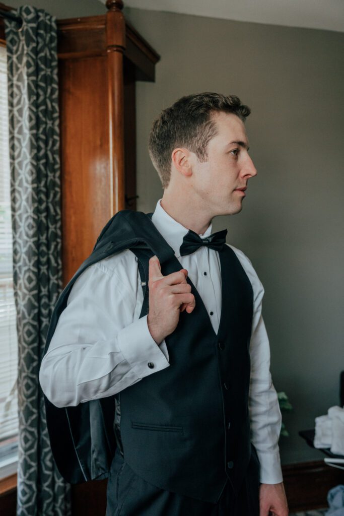 Groom standing in front of window with jacket over his shoulder as he gets ready for romantic Massachusetts wedding.