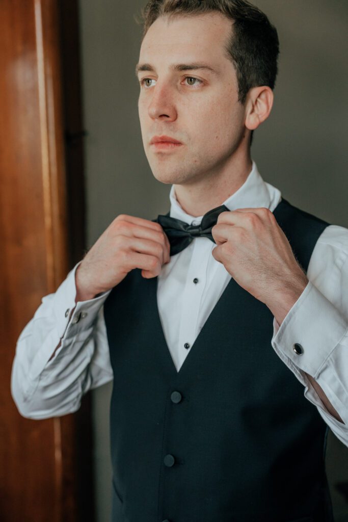 Groom standing in front of window fixing his bowtie for summer Massachusetts wedding.