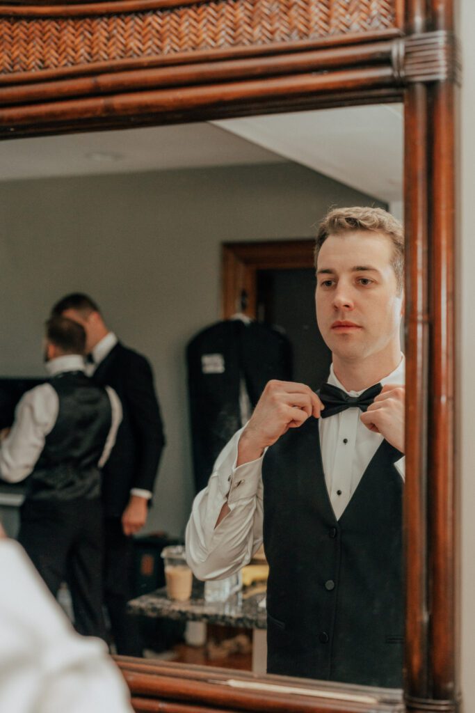 Groom looking in the mirror as he puts on is bowtie for romantic Massachusetts wedding.
