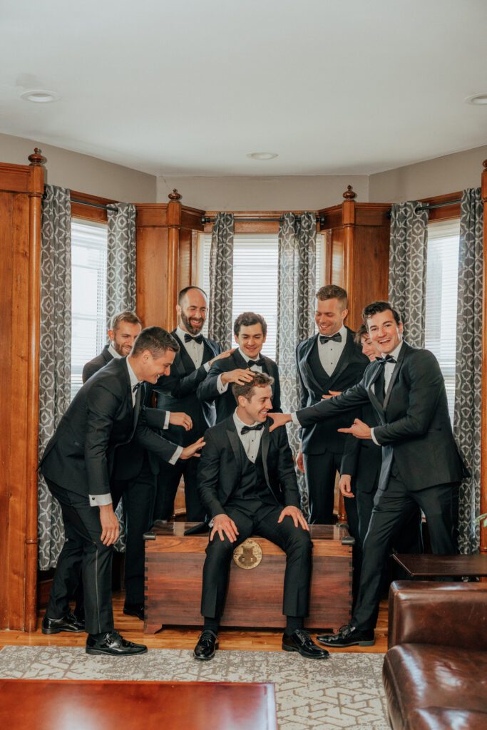 Groom and groomsmen laugh and smile as they sit next to a window in Massachusetts.