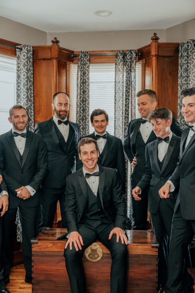 Closeup of groom and groomsmen laugh and smile as they sit next to a window in Massachusetts.