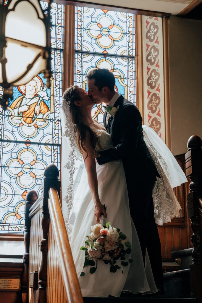 Groom dip kissing a bride in front of stained glass during summer wedding at Quinet Saint John Garden of Our Lady in Massachusetts.