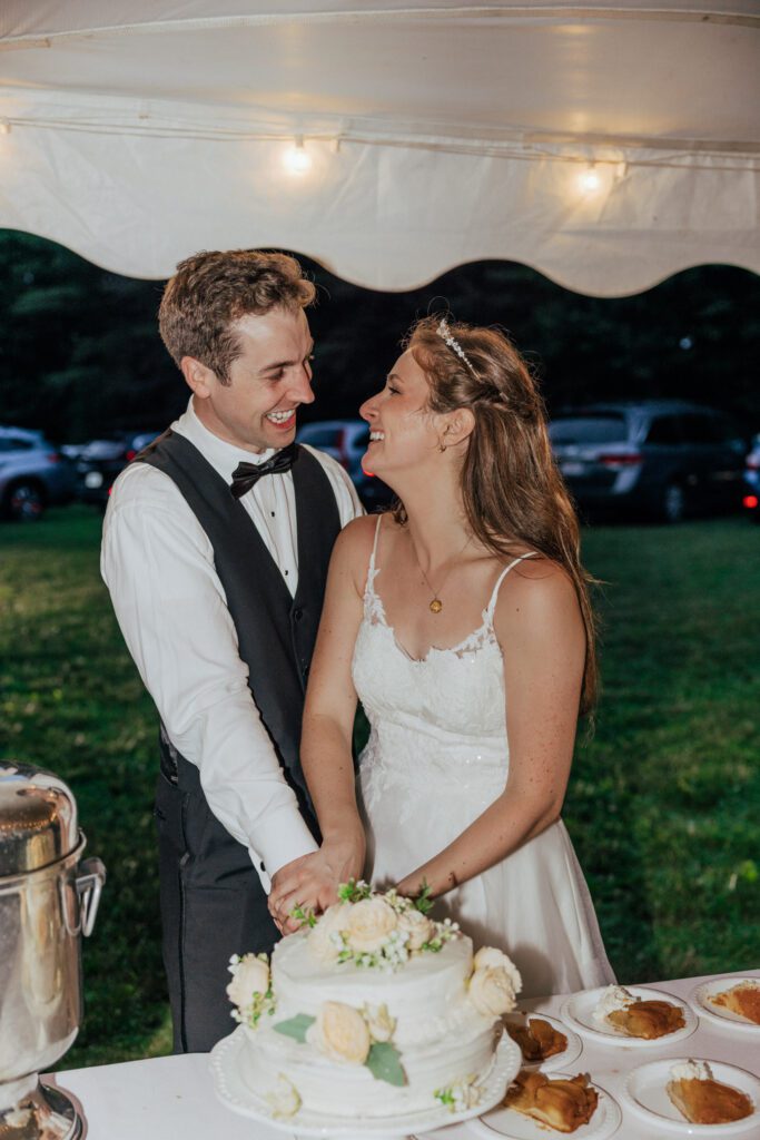 Groom and bride smiling while cutting wedding cake during wedding reception in Massachusetts.