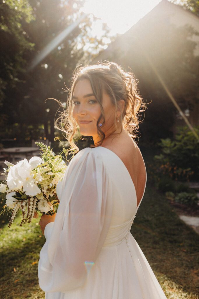Bride romantically looking over her shoulder in garden with the sun shining at New England Botanical Garden at Tower Hill in Boylston, Massachusetts.