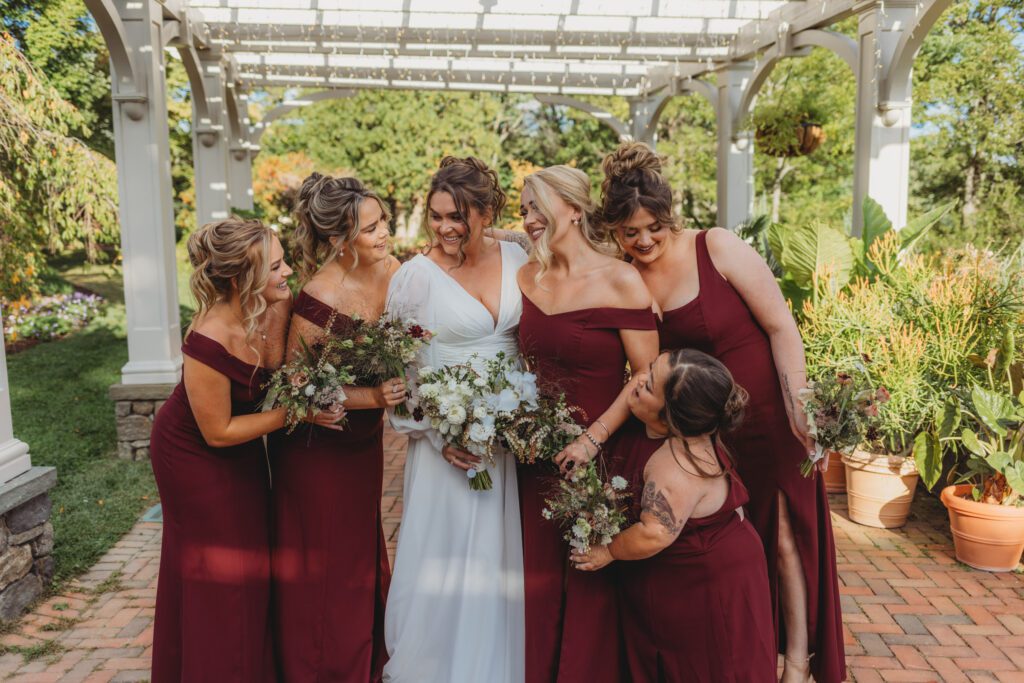 Candid bride and bridesmaids smiling and looking at each other under archway garden at Tower Hill in Boylston, Massachusetts.