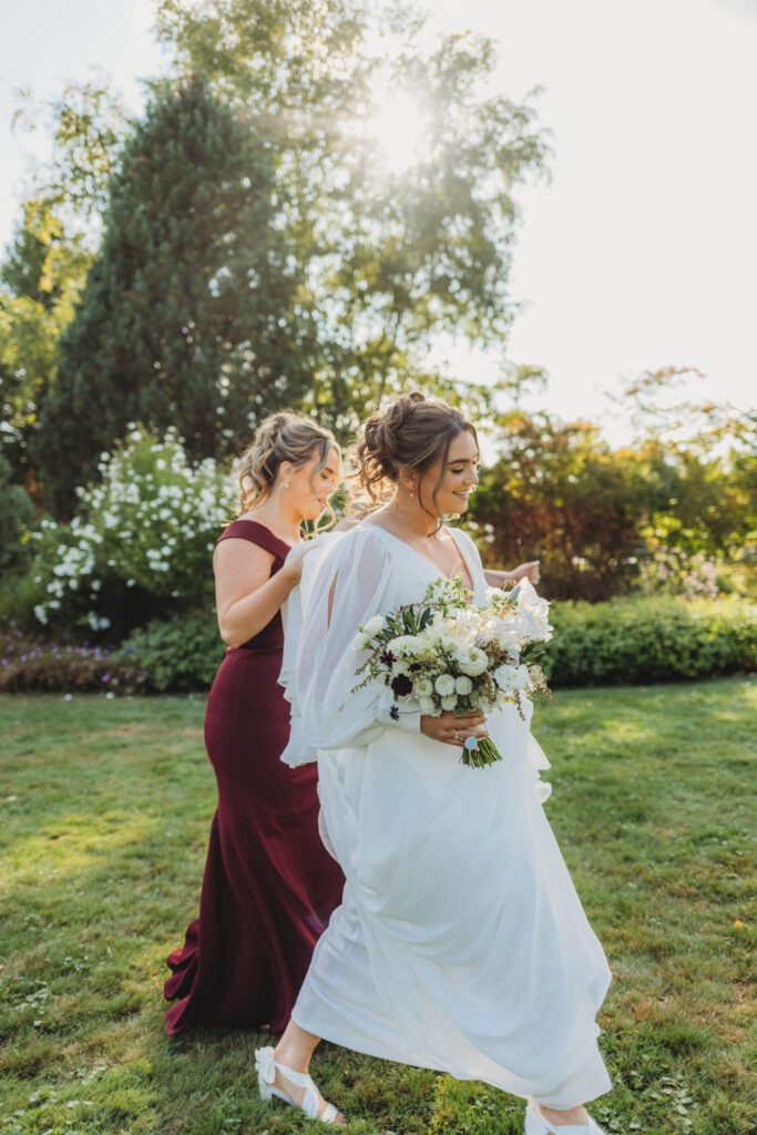 Bride and bridesmaid walking through garden, and holding up the brides wedding dress at New England Botanical Garden at Tower Hill in Boylston, Massachusetts.