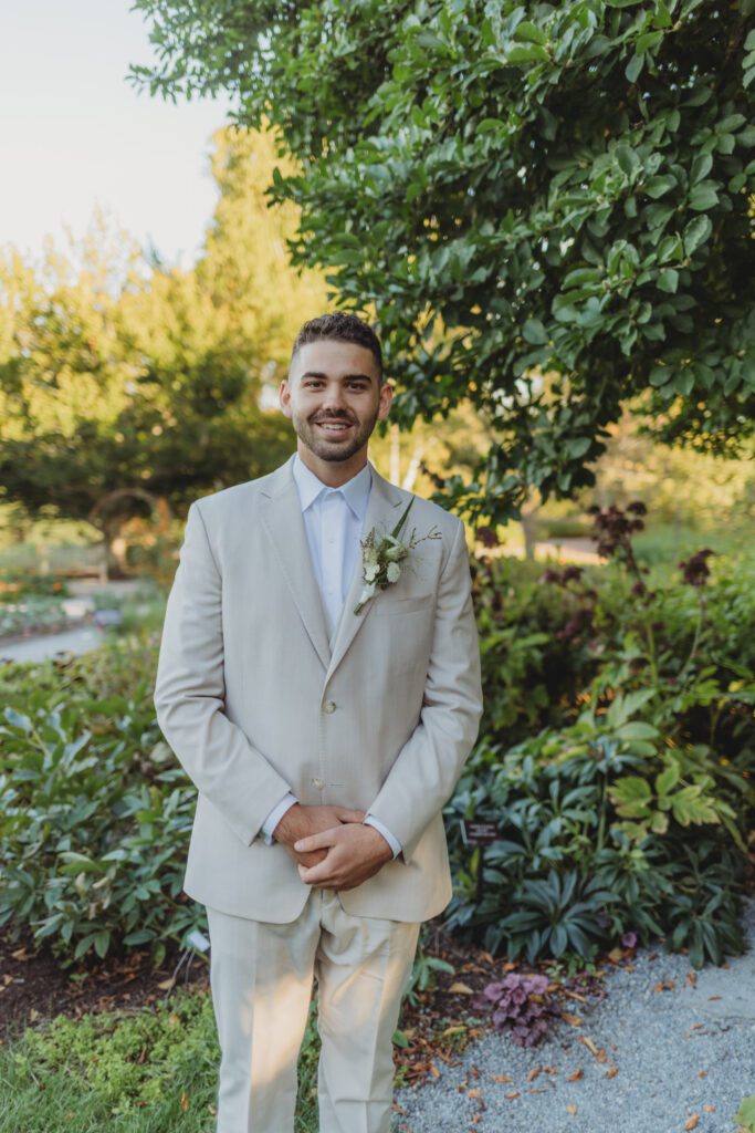 Groom smiling in a tan suit standing in a garden at New England Botanical Garden at Tower Hill in Boylston, Massachusetts.