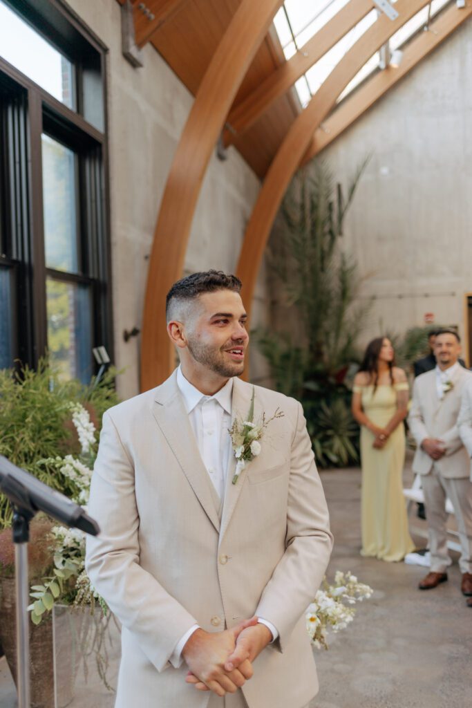 Groom smiling as bride walks down the aisle towards him during greenhouse wedding at Tower Hill in Boylston, Massachusetts.