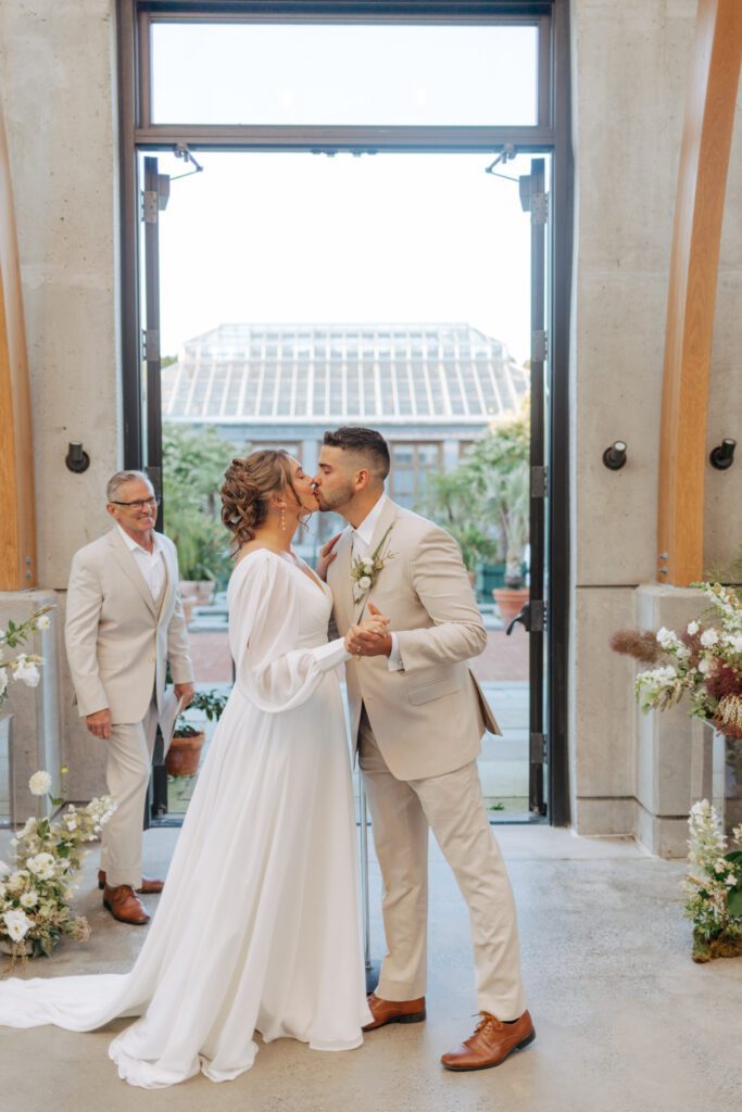 Bride and groom share first kiss during greenhouse wedding ceremony at Tower Hill in Boylston, Massachusetts.