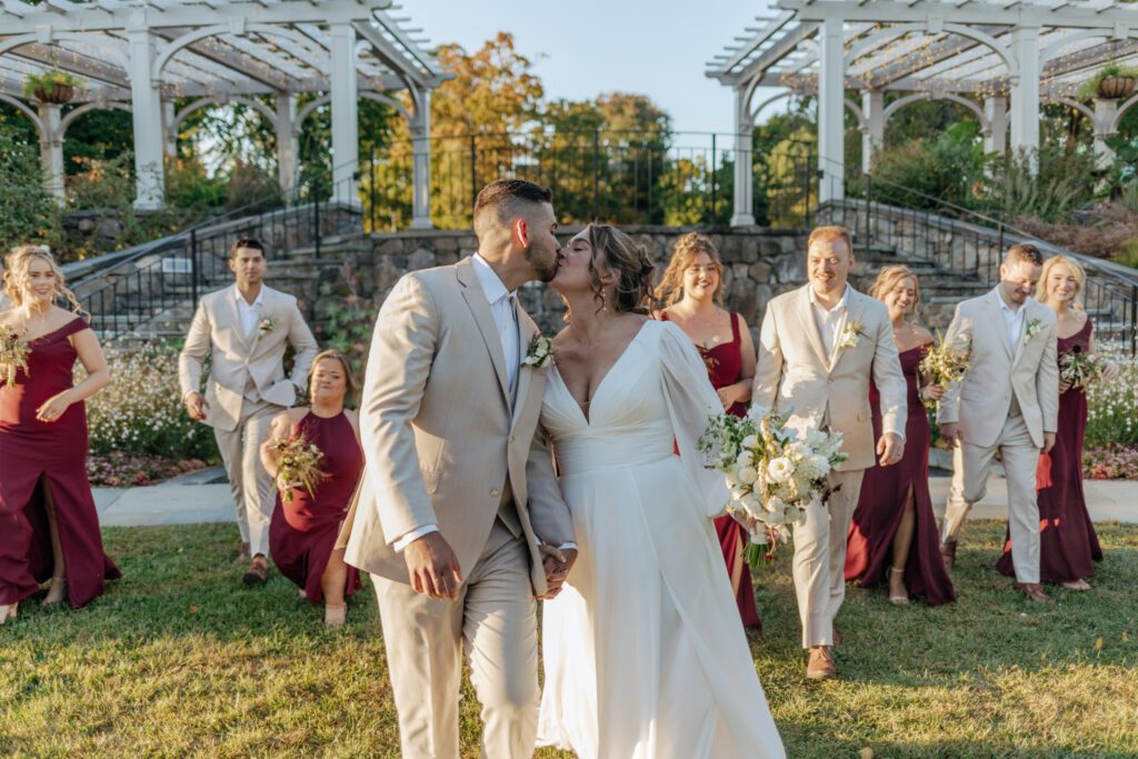 Bride and groom kiss as bridal party walks behind them in front of stone stairs at Tower Hill in Boylston, Massachusetts.