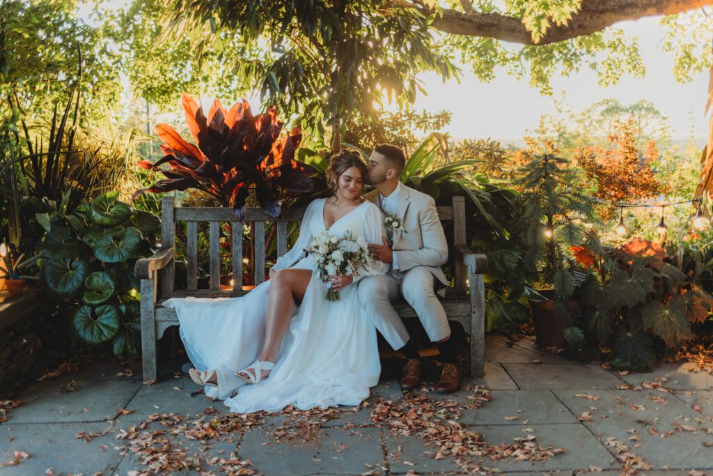 Groom kissing bride on the head as they are sitting on wooden bench in a garden at Tower Hill in Boylston, Massachusetts.