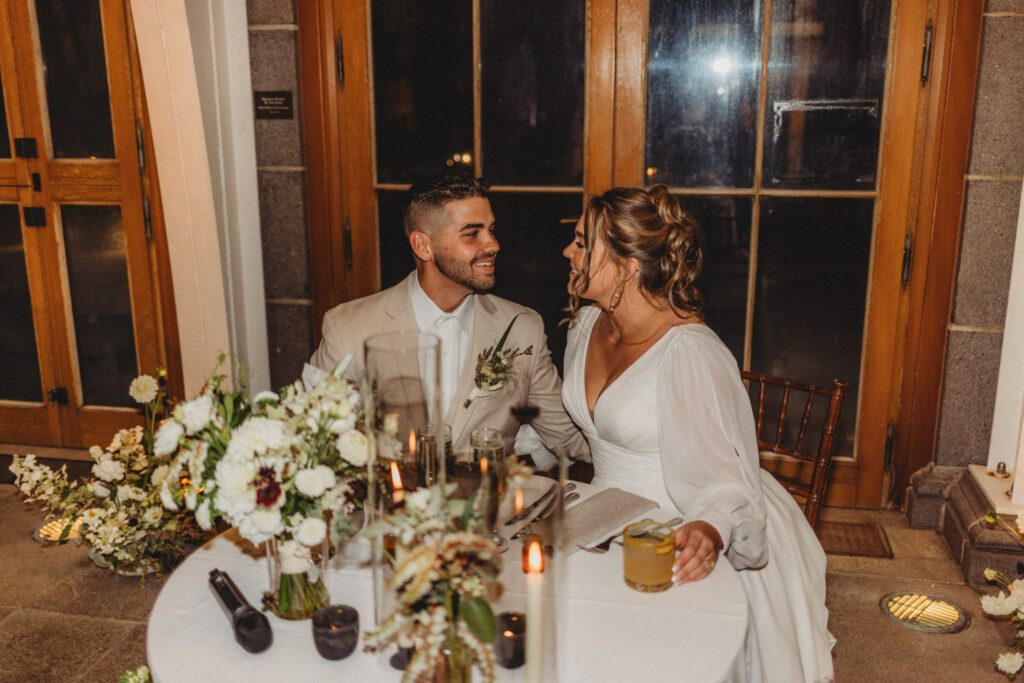 Bride and groom sitting at their sweetheart table smiling at each other at greenhouse reception at Tower Hill in Boylston, Massachusetts.
