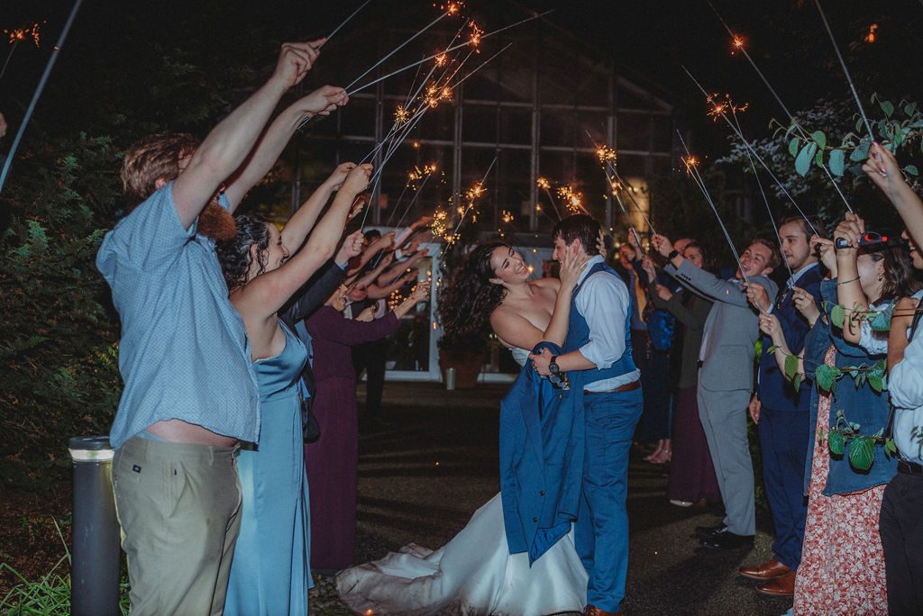 Bride and groom smiling as they exit their wedding with their guest holding sparklers at Roger Williams Botanical Garden Park in Providence, Rhode Island.