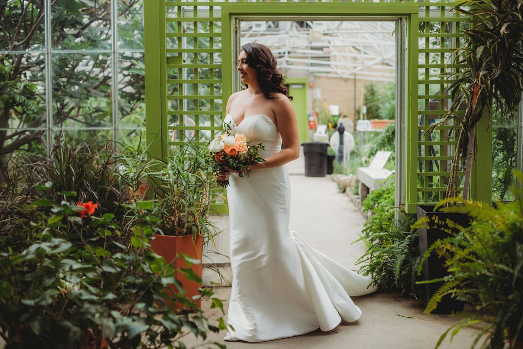 Bride walking through a greenhouse holding flowers and smiling at Roger Williams Botanical Garden Park in Providence, Rhode Island.