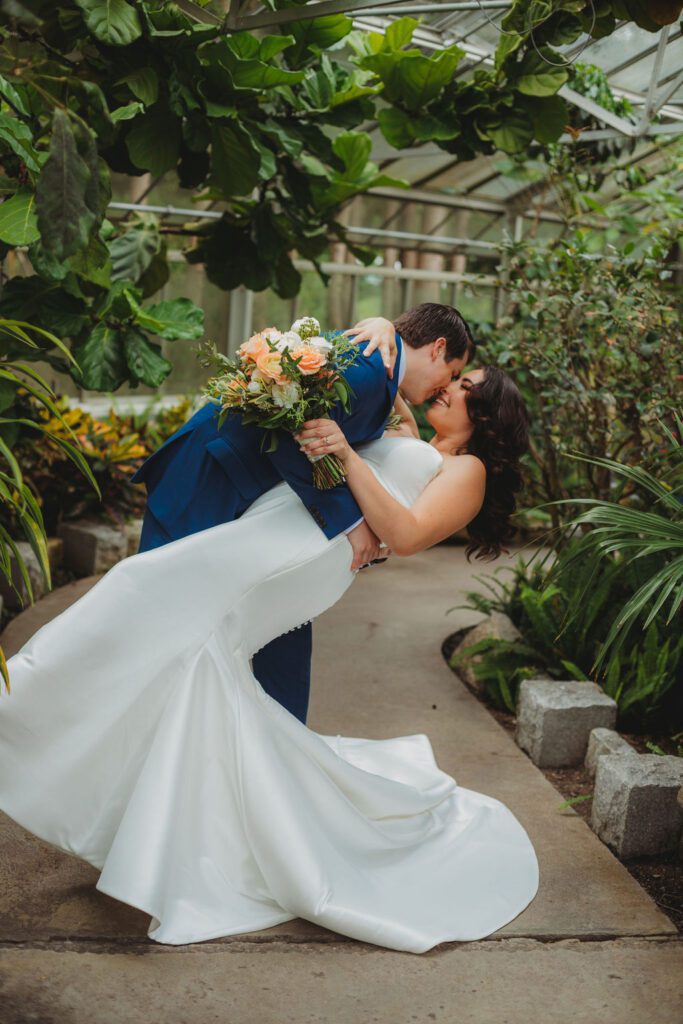 Groom dips bride and kisses her during first look in greenhouse at Roger Williams Botanical Garden Park in Providence, Rhode Island.