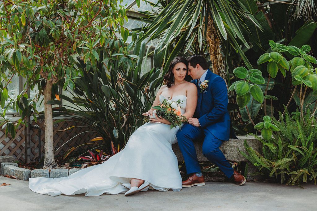 Bride and groom sitting on stone bench as groom leans into bride romantically in greenhouse at Roger Williams Botanical Garden Park in Providence, Rhode Island.
