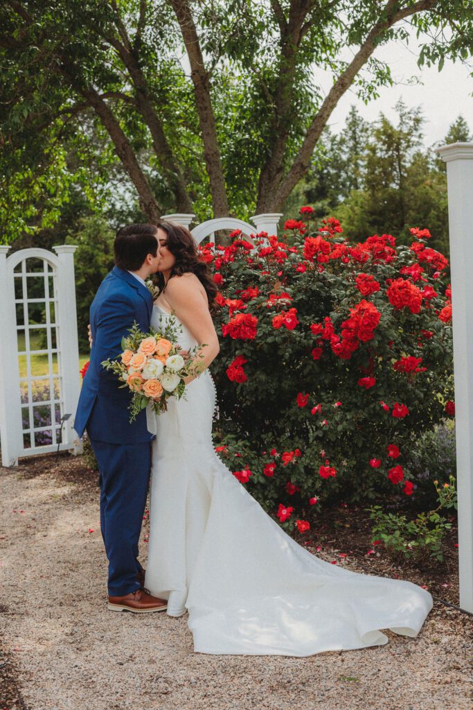 Bride and groom romantically kiss in front of a rose bush at Roger Williams Botanical Garden Park in Providence, Rhode Island.