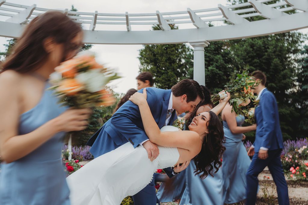 Groom dipping bride as bridal party walks by them in a blurry way in rose garden at Roger Williams Botanical Garden Park in Providence, Rhode Island.
