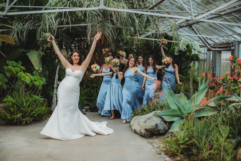 Bride walking into greenhouse with her arms in the arm followed by her bridesmaid at Roger Williams Botanical Garden Park in Providence, Rhode Island.