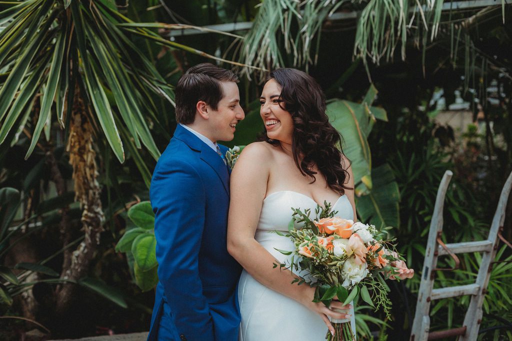 Bride and groom smiling romantically as bride looks over her shoulder in a greenhouse at Roger Williams Botanical Garden Park in Providence, Rhode Island.