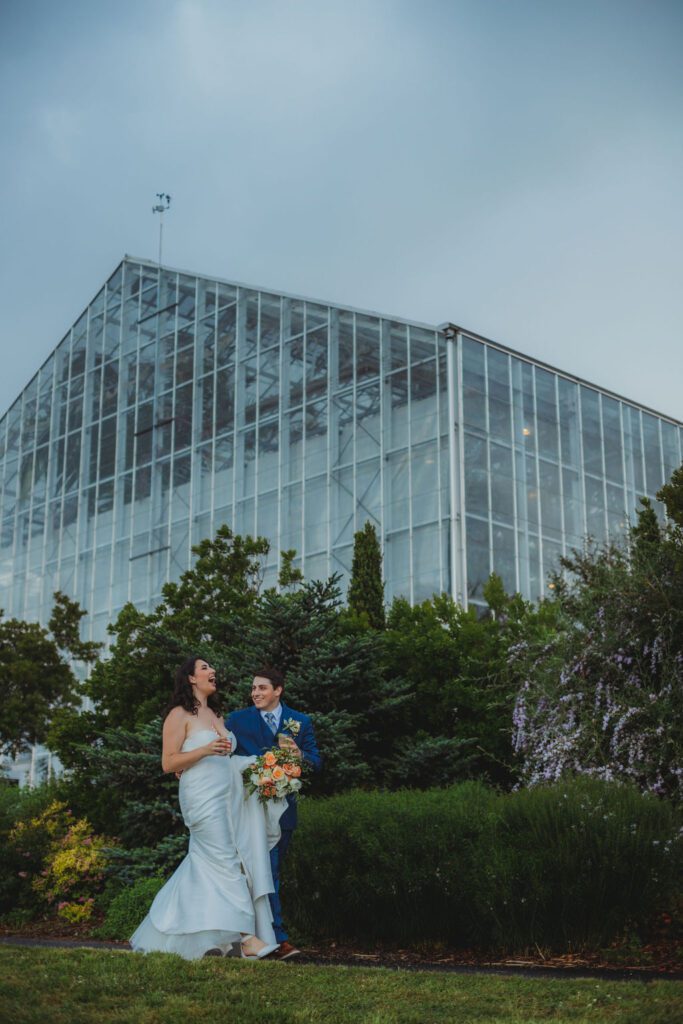 Bride and groom laughing and walking outside a greenhouse at Roger Williams Botanical Garden Park in Providence, Rhode Island.