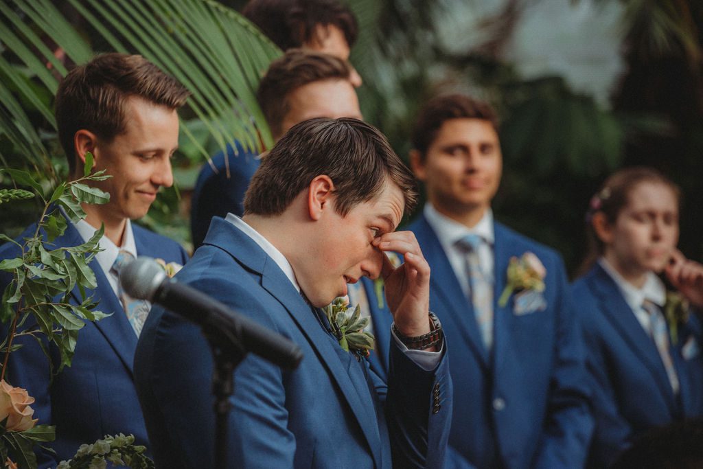 Groom wiping away his tear seeing bride walk down the aisle in greenhouse at Roger Williams Botanical Garden Park in Providence, Rhode Island.