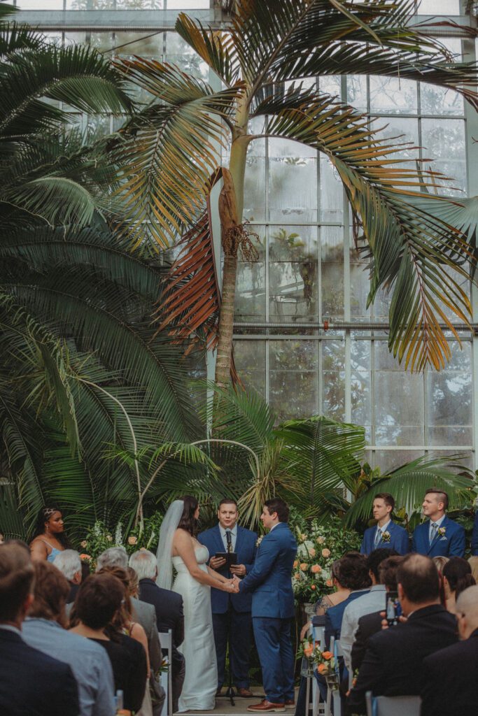 Bride and groom holding hands during wedding ceremony in greenhouse at Roger Williams Botanical Garden Park in Providence, Rhode Island.