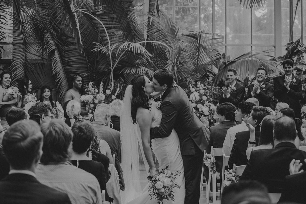 Groom dipping bride in the middle of the aisle and kissing after wedding ceremony in greenhouse at Roger Williams Botanical Garden Park in Providence, Rhode Island.