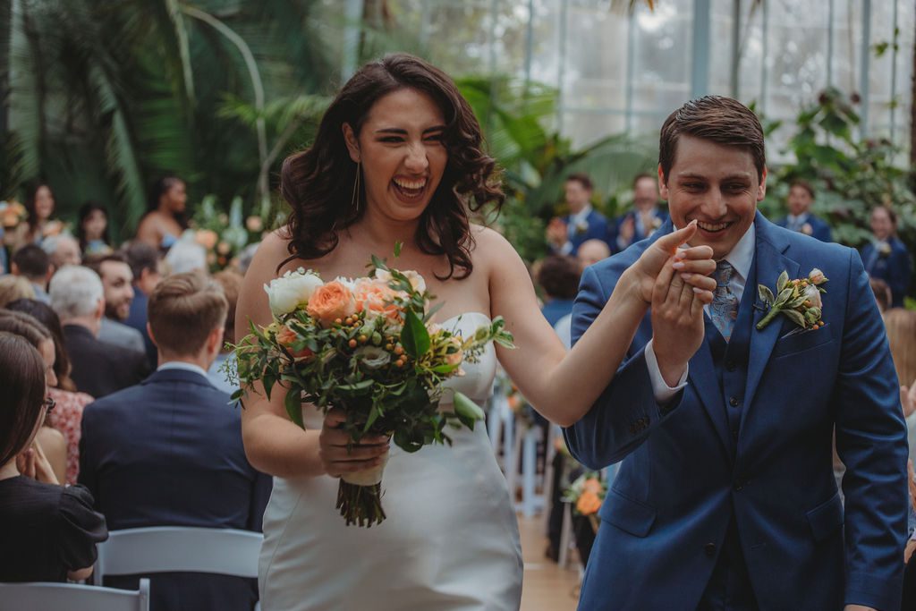 Bride and groom smiling and exiting greenhouse ceremony at Roger Williams Botanical Garden Park in Providence, Rhode Island.