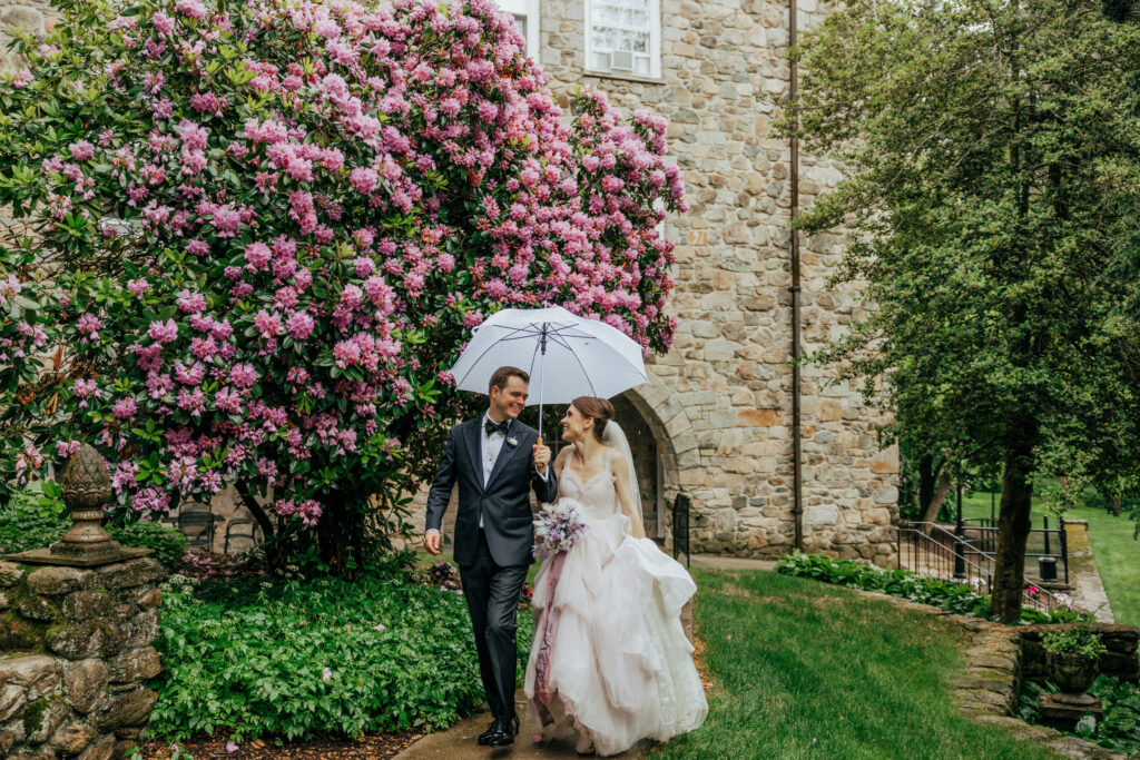 Bride and groom walking as they laugh under clear umbrella in front of stone building at The Conner Center in Dover, Massachusetts.