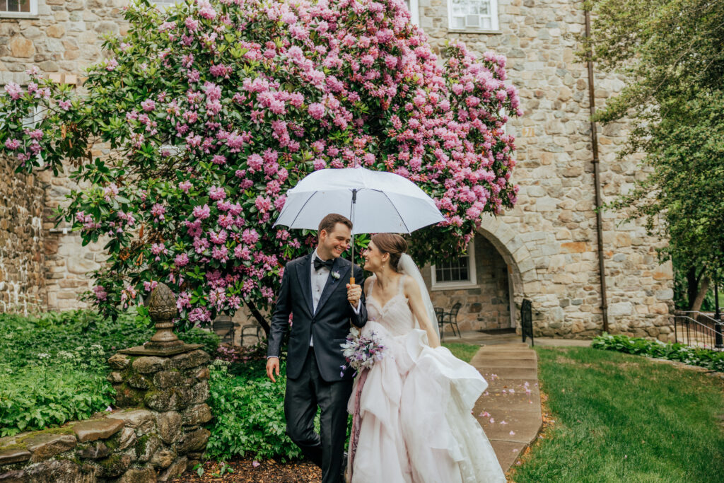 Bride and groom walking under clear umbrella in front of stone building and pink flowers at The Conner Center in Dover, Massachusetts.