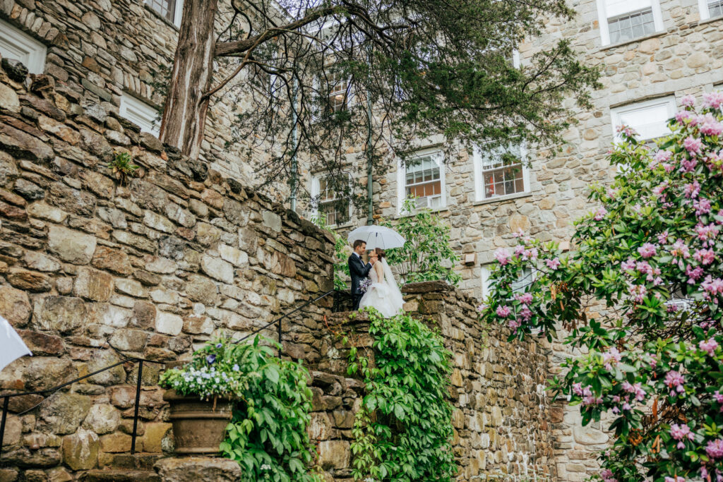 Romantic bride and groom standing on stone stair case under clear umbrella at The Conner Center in Dover, Massachusetts.
