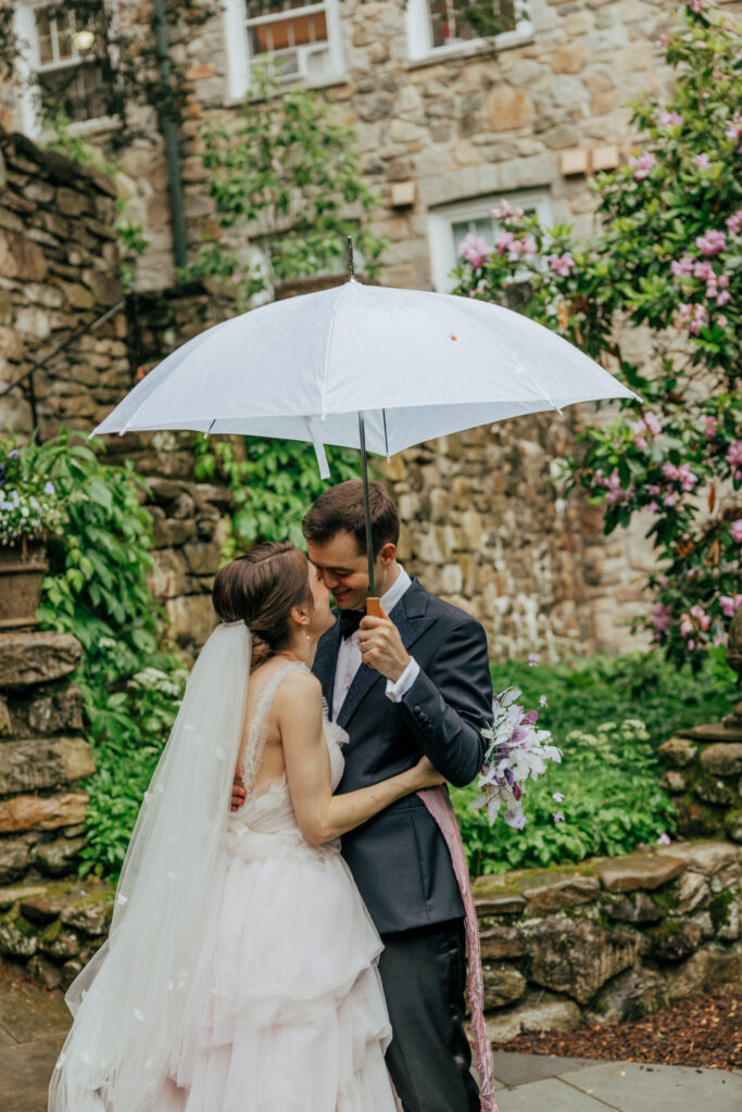 Bride and groom smiling at each other under clear umbrella in front of stone building at The Conner Center in Dover, Massachusetts.