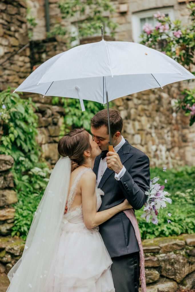 Bride and groom kissing under clear umbrella in front of stone building at The Conner Center in Dover, Massachusetts.